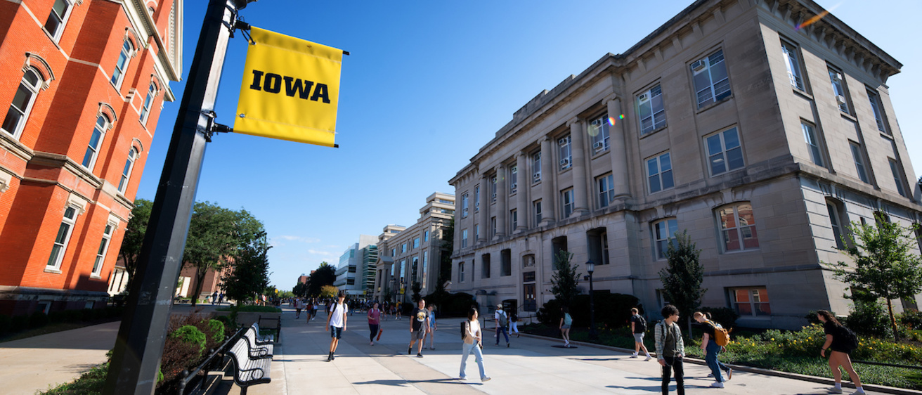 T. Anne Cleary walkway from the corner of Calvin Hall, facing Gilmore hall. A lamppost with an Iowa banner is in the foreground.