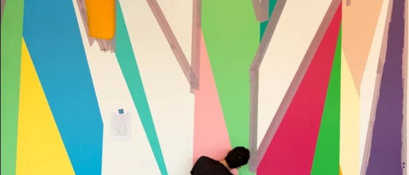 A man in black clothing helps paint a colorful geometric mural in the Stanley Museum of Art